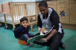 Little boy sitting down with a healthcare professional crouching next to them reading him a picture book.