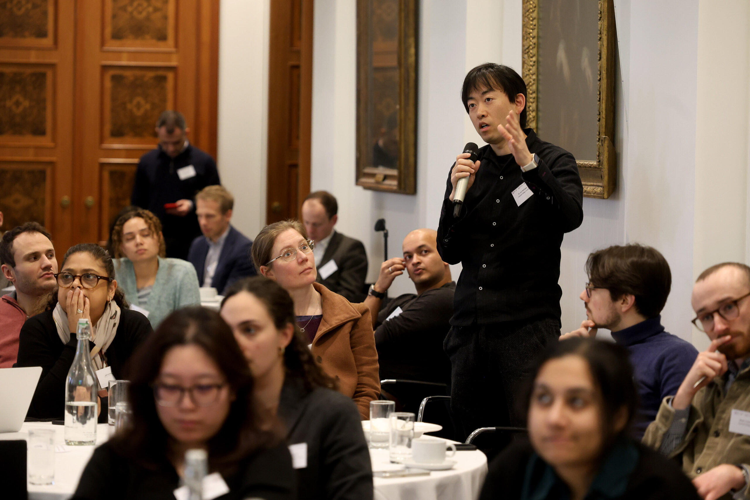 Conference attendees sitting at round tables. A man is standing holding a microphone to ask a question.