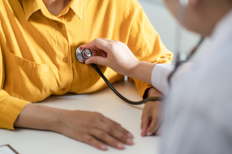 A doctor in a white coat holding a stethoscope on the chest of a patient wearing a yellow shirt.