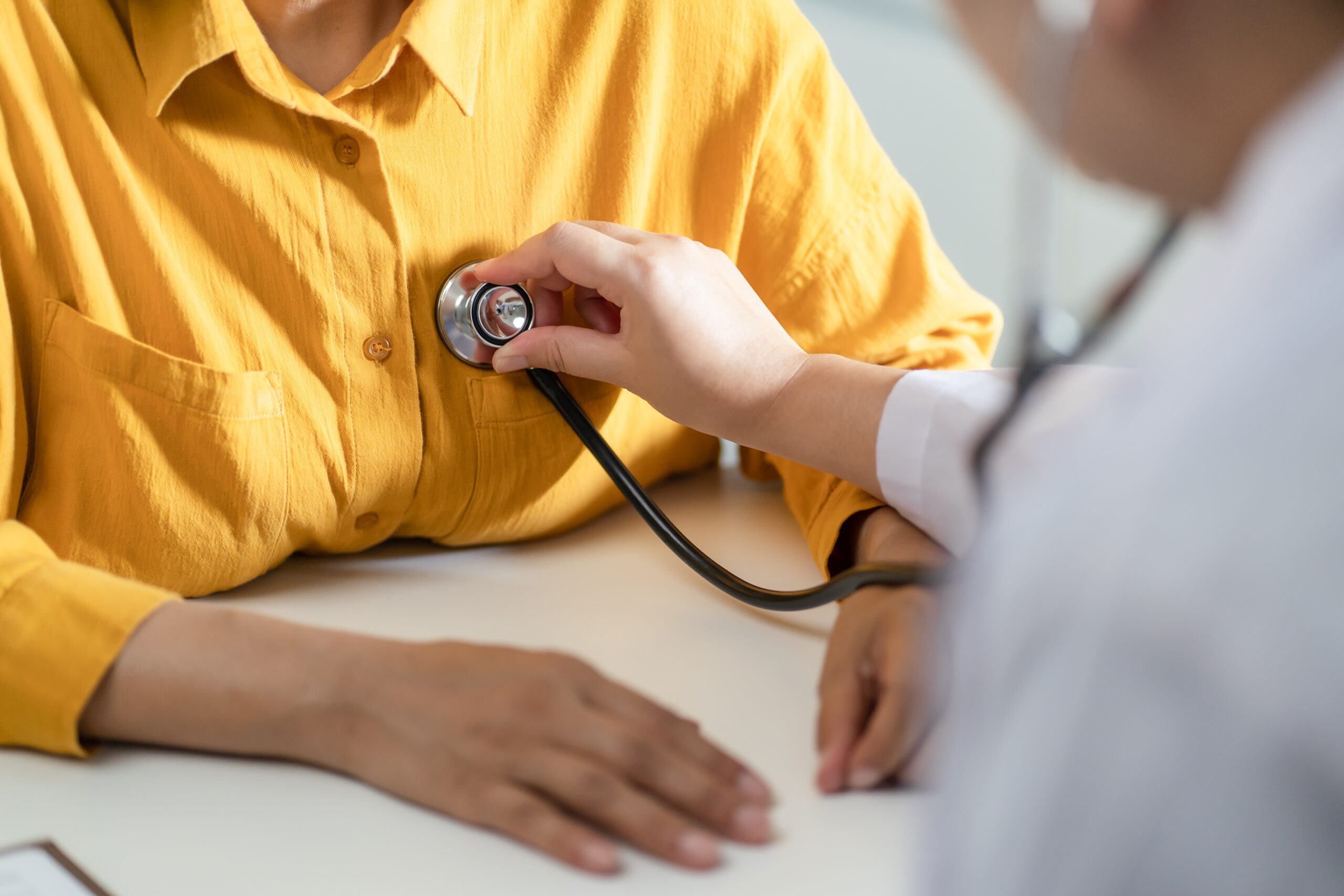 A doctor in a white coat holding a stethoscope on the chest of a patient wearing a yellow shirt.